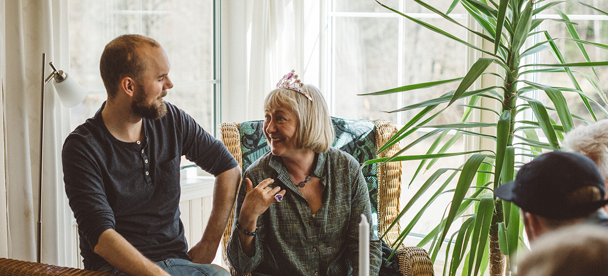 Ein junger Mann und eine ältere Frau mit Geburtstagskrone auf dem Kopf unterhalten sich in einem Wohnzimmer