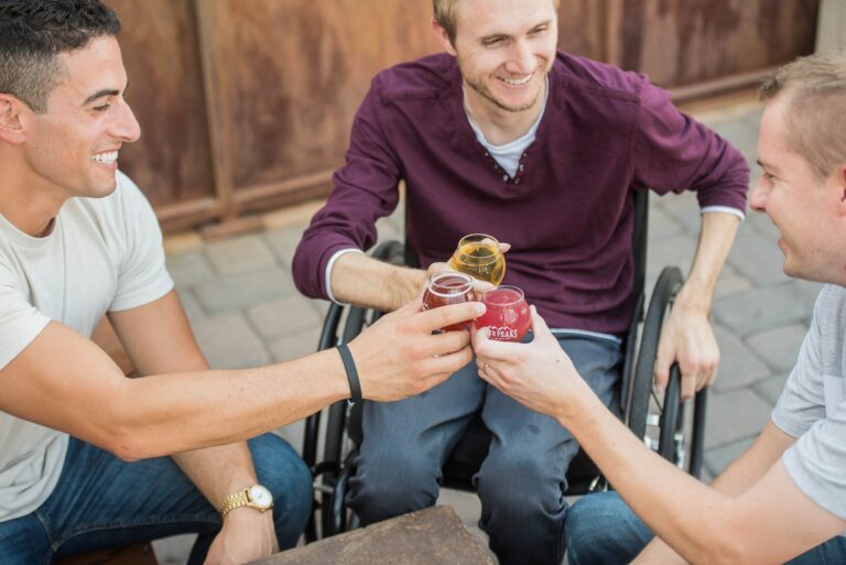 Drei junge Männer, einer davon im Rollstuhl, stoßen mit Getränken an.