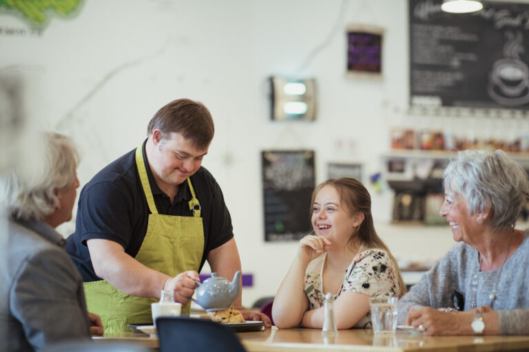 Person mit Downsyndrom serviert anderen Tee und Kekse am Tisch.