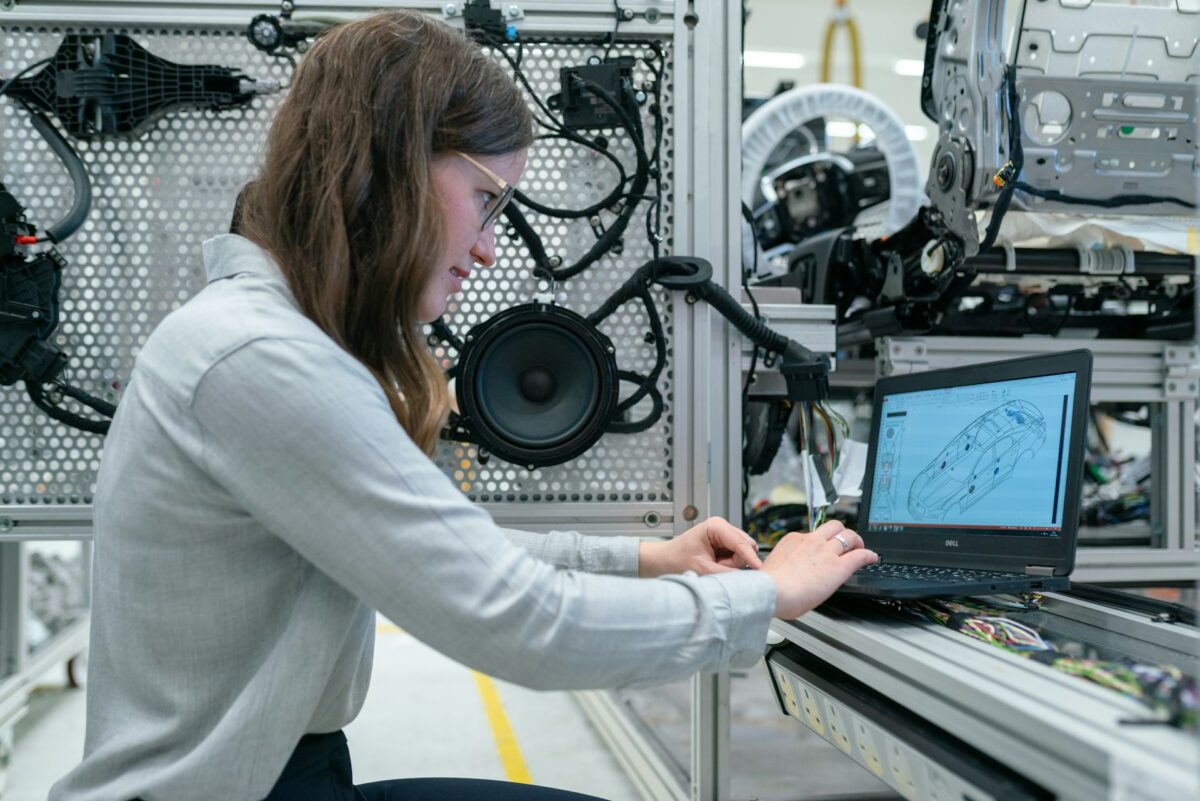 Person arbeitet in einer technischen Entwicklungsumgebung an einem Laptop mit CAD-Modell eines Fahrzeugs; im Hintergrund sind Fahrzeugkomponenten und Kabelstränge zu sehen.